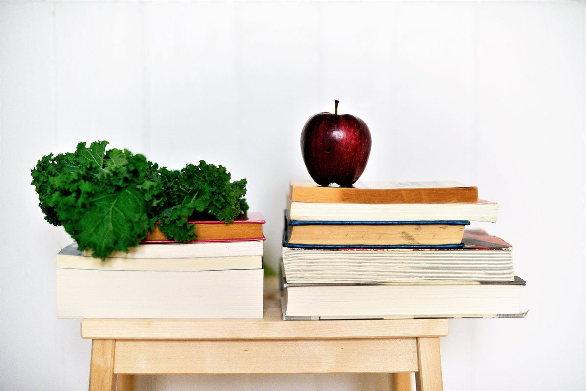 A wooden stool holds two stacks of books. On the left pile, a bunch of green kale rests, while on the right pile, a red apple sits on top. The background is a plain white wall, creating a simple and clean composition.