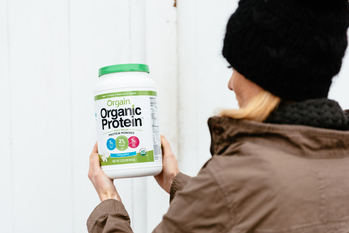 A person wearing a black hat and brown jacket holds a large container of Orgain Organic Protein powder. The container is white with a green lid and blue, red, and green labels advertising 21 grams of protein and additional nutritional information.