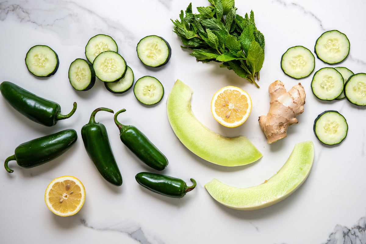 A flat lay of fresh ingredients on a white marble surface, including sliced cucumbers, whole jalape√±os, melon slices, a halved lemon, a chunk of ginger root, and a bunch of mint leaves.