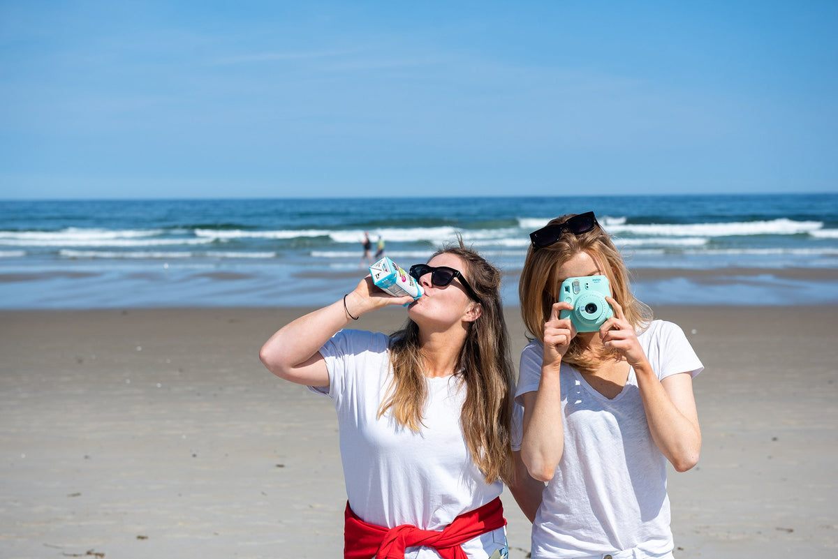 Two women stand on a sandy beach with waves in the background. The woman on the left drinks from a can while the woman on the right uses a mint-colored camera. Both wear sunglasses, casual white shirts, and appear to be enjoying a sunny day.