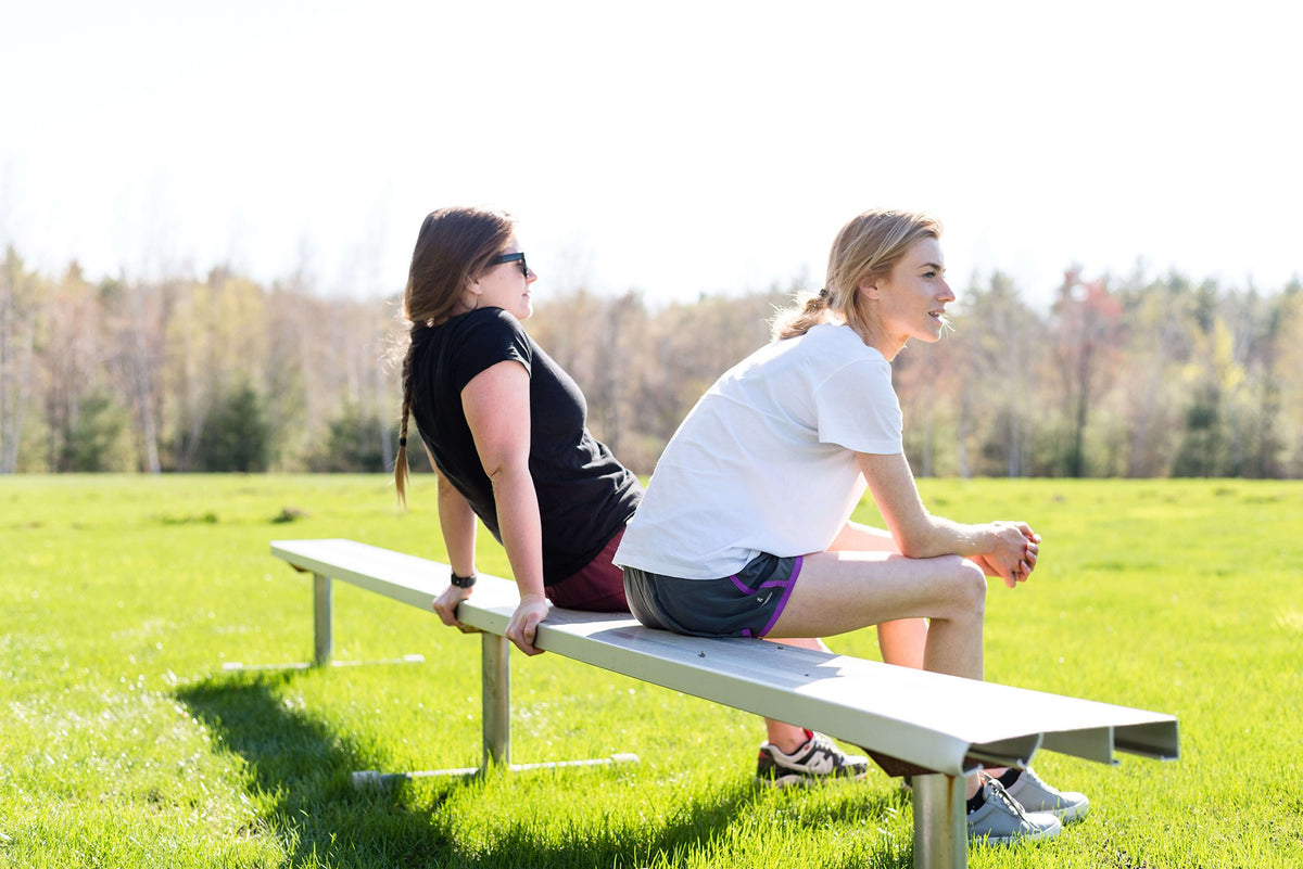 Two women sitting on a bench in a grassy field under a bright sky, with trees in the background.