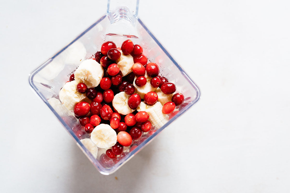 Overhead view of a blender jar containing chopped bananas and vibrant red cranberries, placed on a white surface. The mixture appears fresh and ready for blending.