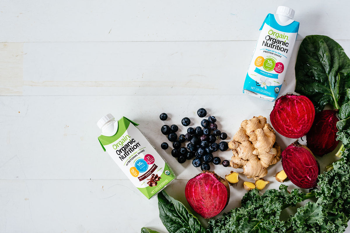An overhead shot of two Orgain Organic Nutrition drinks in cartons, one green and one blue, placed on a light wooden surface. Surrounding the drinks are fresh vegetables and fruits, including beetroots, kale, spinach, blueberries, and ginger.