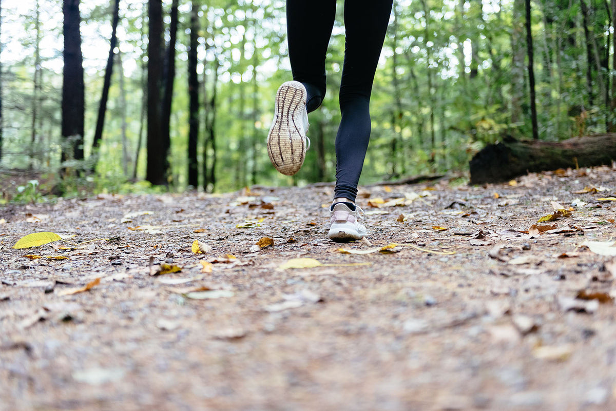 A person running on a forest trail, with their legs and feet visible. The path is covered in fallen leaves, and trees with green foliage surround the scene. The image captures motion and the tranquility of nature.