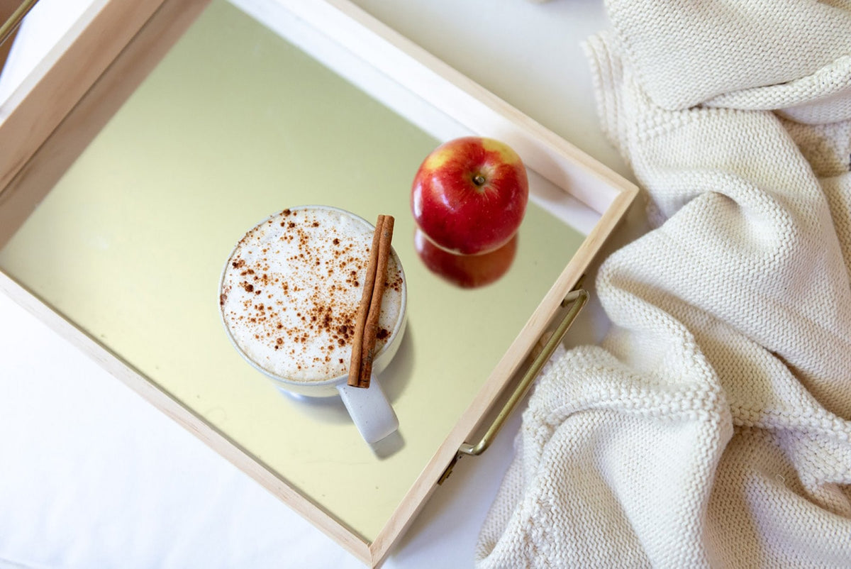 A wooden tray holds a foamy, cinnamon-topped latte in a clear glass mug, accompanied by a small spoon. Beside it, there's a shiny red apple. The tray sits on a light-colored knitted blanket, adding a cozy touch to the scene.