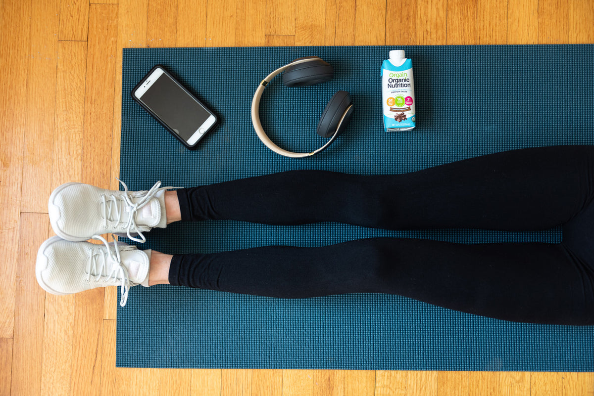 A person in black leggings and white sneakers lies on a blue yoga mat. Near their legs are a smartphone, wireless headphones, and a bottle of protein shake placed on a wooden floor.