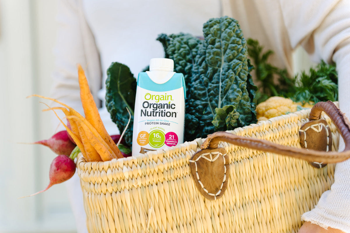 A person carries a woven basket filled with fresh vegetables, including carrots and leafy greens, along with a carton of Orgain Organic Nutrition protein shake. The basket has brown handles and intricate detailing.