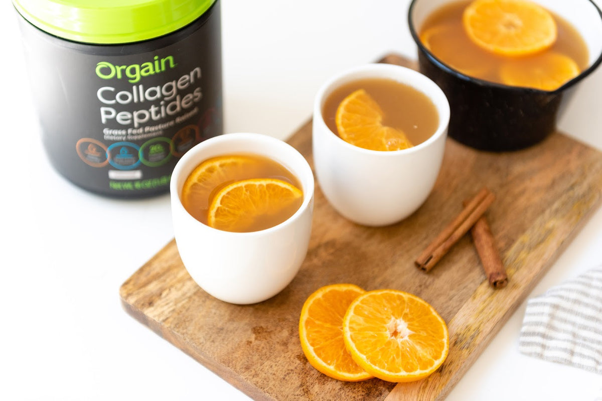 Two cups with light brown liquid on a wooden board with orange slices and cinnamon. Orgain Collagen Peptides in the background.