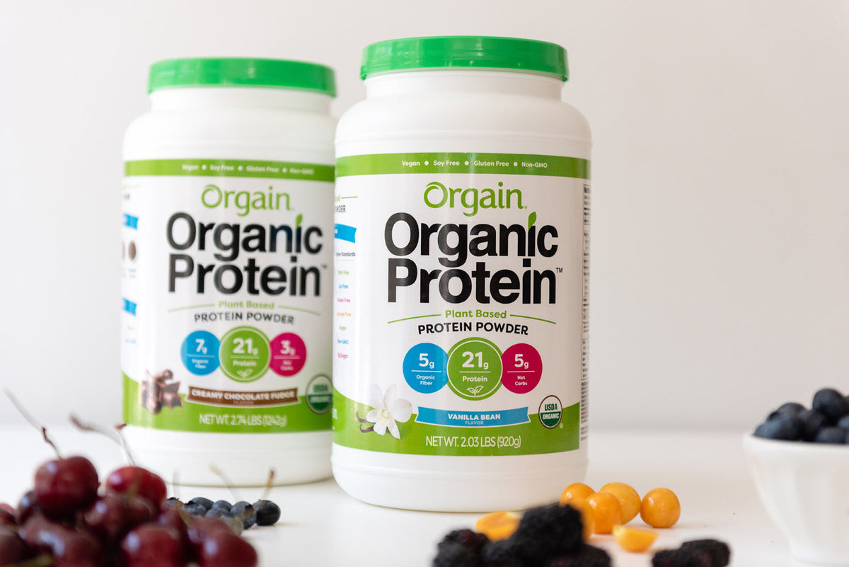 Two containers of Orgain Organic Protein plant-based protein powder are displayed. The container in the foreground is vanilla bean flavor, and the one in the background is creamy chocolate fudge flavor. Fresh berries and cherries are scattered around.