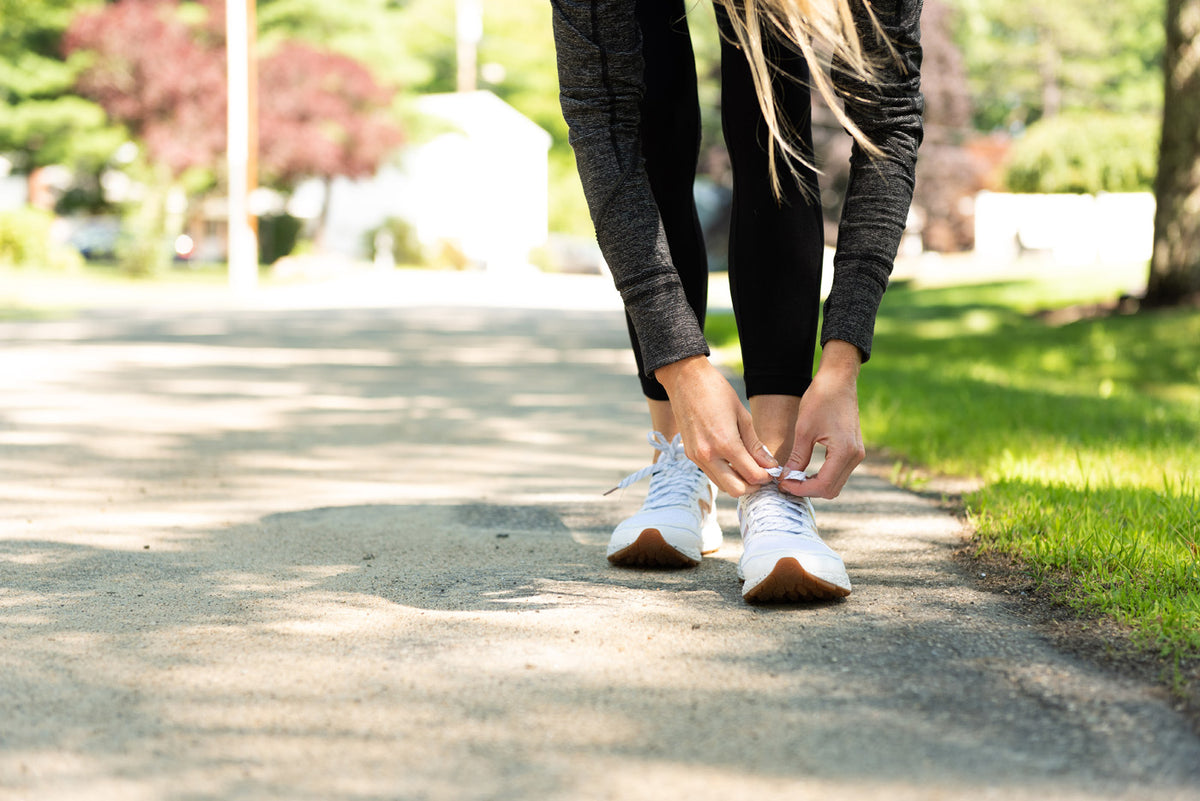 A person with long hair, dressed in a dark long-sleeve top and black leggings, bends down to tie the laces of their white sneakers on a sunny pathway. Green grass and trees are visible in the background.