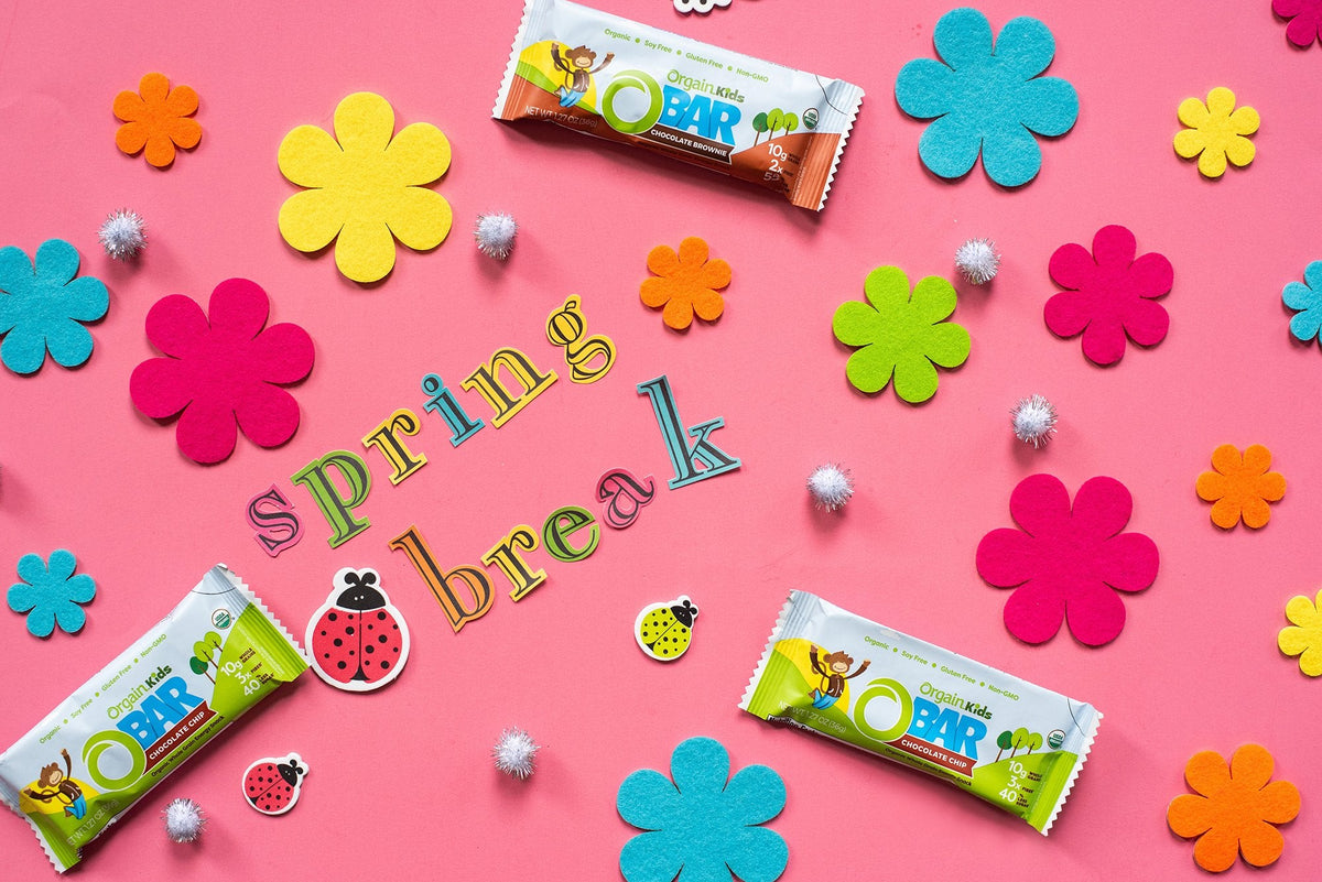 A festive arrangement on a bright pink background displays colorful felt flowers, ladybug decorations, and foam letters spelling "spring break." Three organic kids' O Bar granola bars are placed among the decorations.
