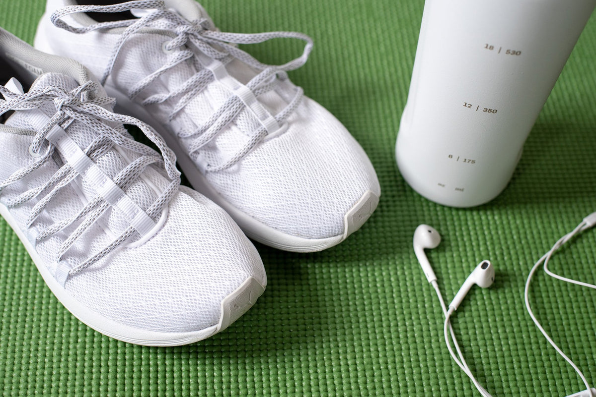 A pair of white athletic shoes are placed next to a white water bottle and a set of white earbuds on a green exercise mat.