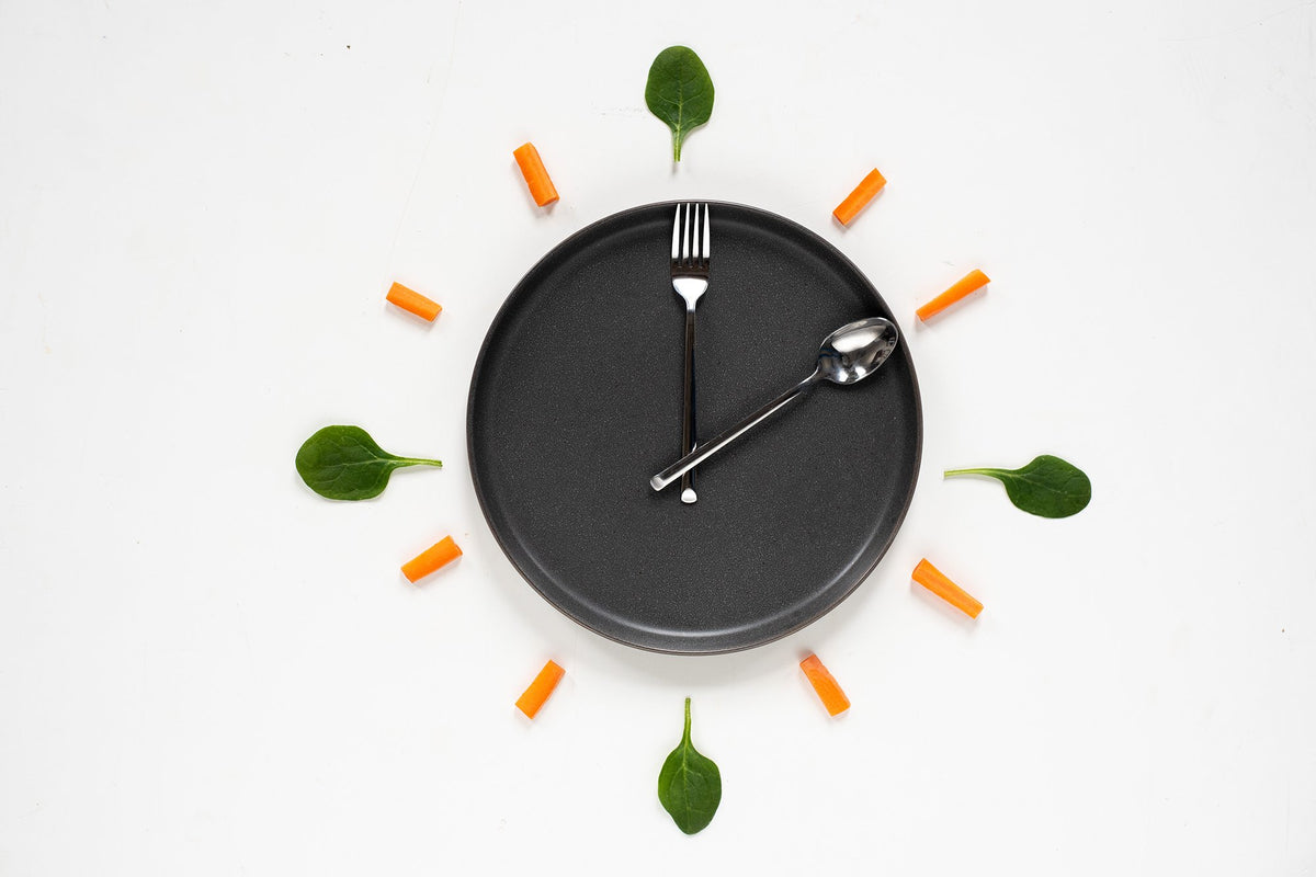 A gray plate on a white surface is arranged to resemble a clock, with a spoon and fork positioned as clock hands. The plate is surrounded by spinach leaves and carrot sticks, acting as hour markers.