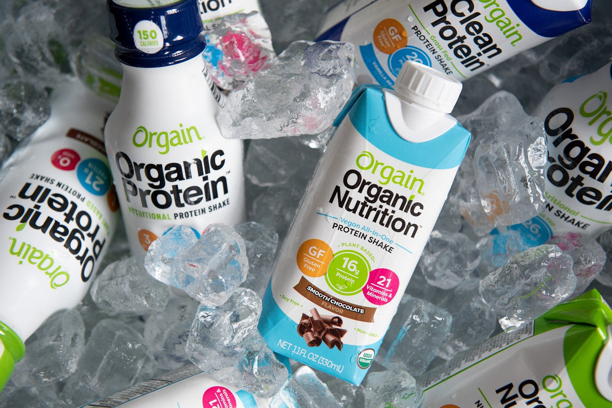 Various Orgain drink products, including Organic Protein, Organic Nutrition, and Orgain Clean Protein, are packed in ice. The labels highlight features like gluten-free, high protein content, and multiple vitamins. The ice keeps the drinks chilled.
