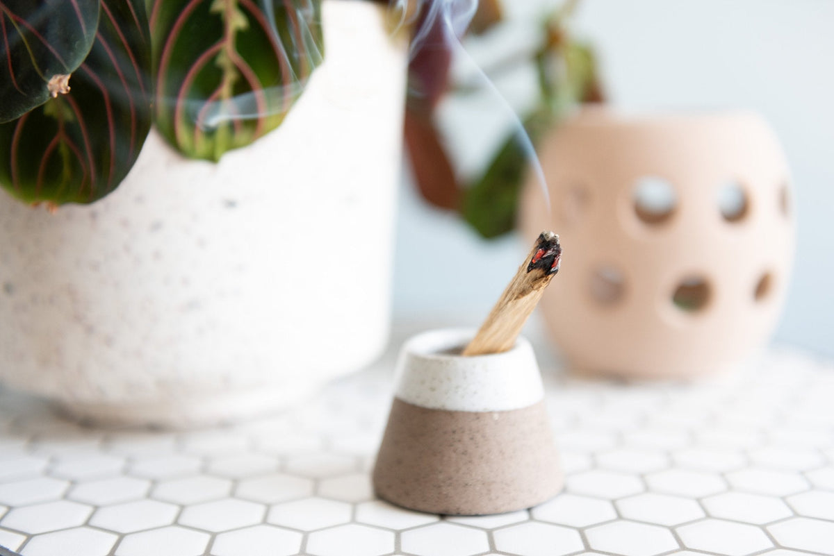Close-up of a lit incense stick emitting smoke, placed in a small, two-toned holder on a white hexagonal-tiled surface. In the background, there are potted plants and a candle holder with circular cutouts.