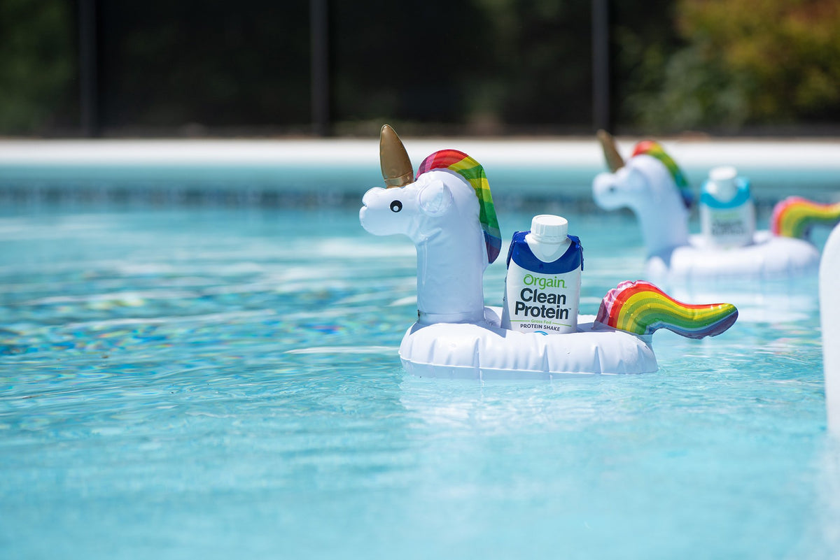 Floating in a pool are inflatable unicorn-shaped drink holders. Each unicorn holds a carton of Orgain Clean Protein. The clear blue water surrounds the unicorn floats, and trees in the background are slightly out of focus.