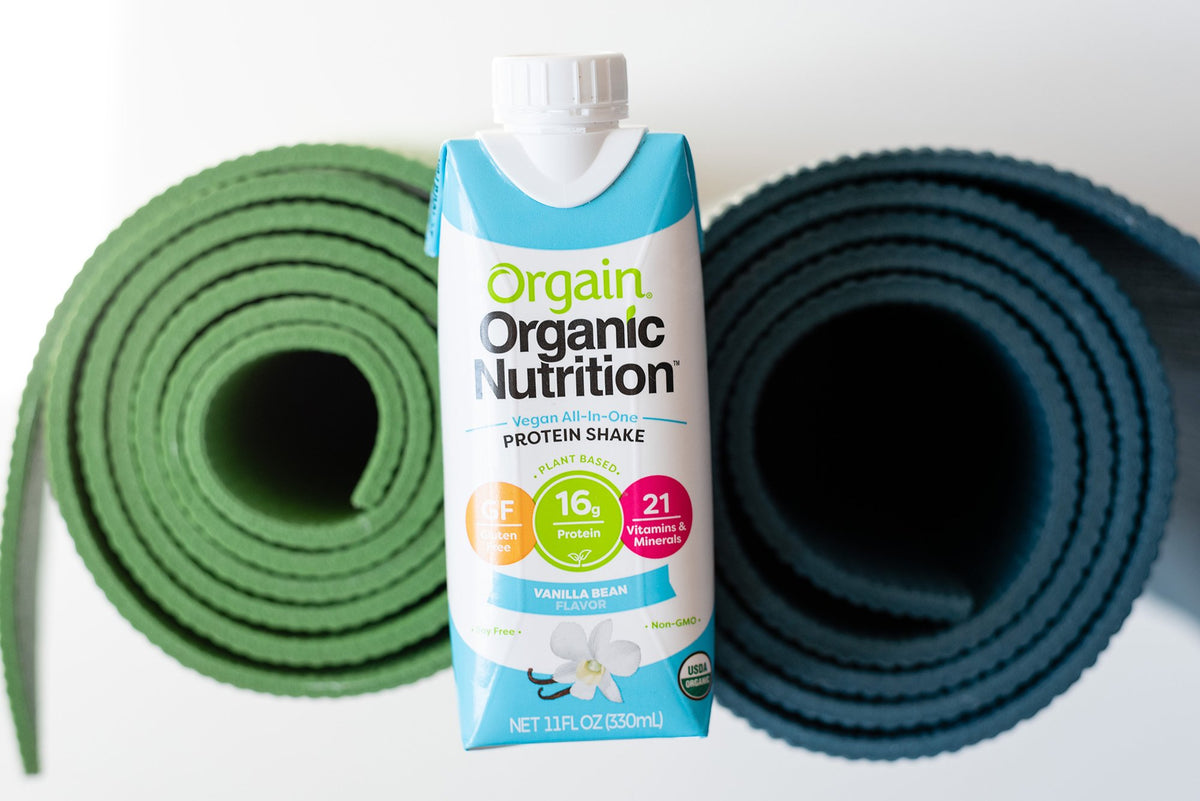 A carton of Orgain Organic Nutrition Vanilla Bean protein shake is placed between two rolled-up yoga mats, one green and one dark blue, against a white background. The protein shake carton displays key nutritional information and benefits.