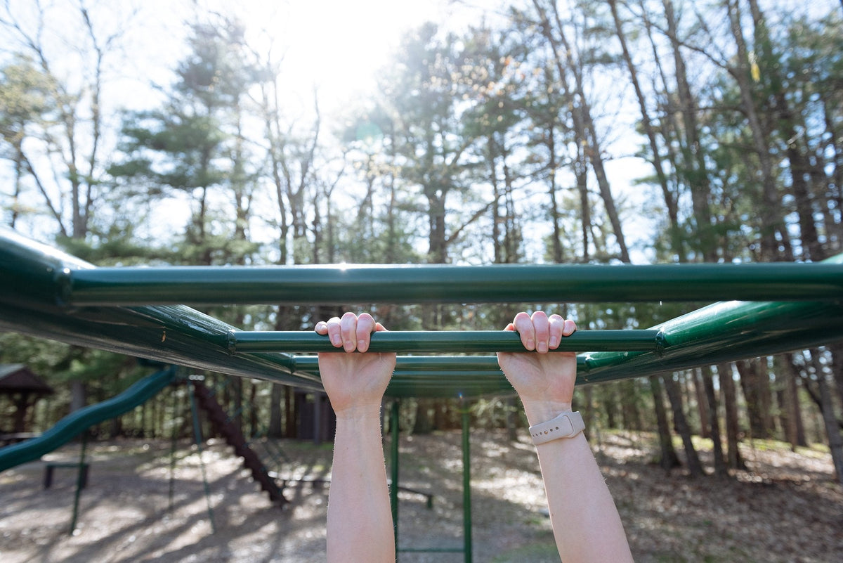 Close-up of hands gripping green monkey bars in an outdoor playground, with a sunlit forest in the background. The person is wearing a beige watch, and there is playground equipment visible in the distance among the trees.