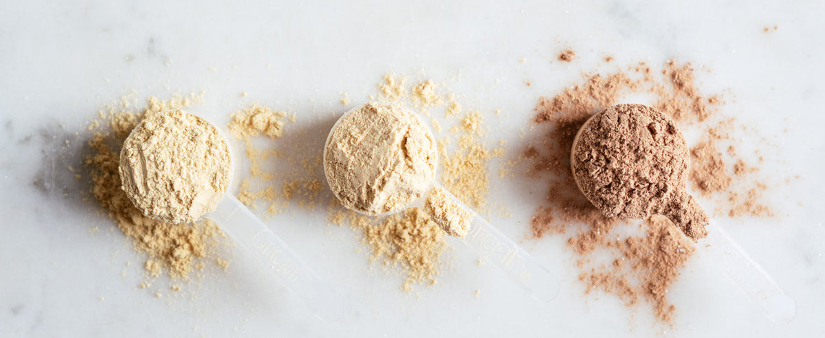 Three scoops of different protein powders, each with a bit of powder spilled around them, on a white marble surface. From left to right: one light yellow, one beige, and one brown. Each scoop has a transparent handle.