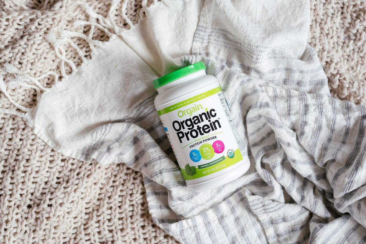 A container of Orgain Organic Protein powder with a green lid rests on a soft, light-colored knit blanket and a white cloth with light gray stripes. The protein powder label highlights 21g protein, 5g fiber, and 0g sugar.