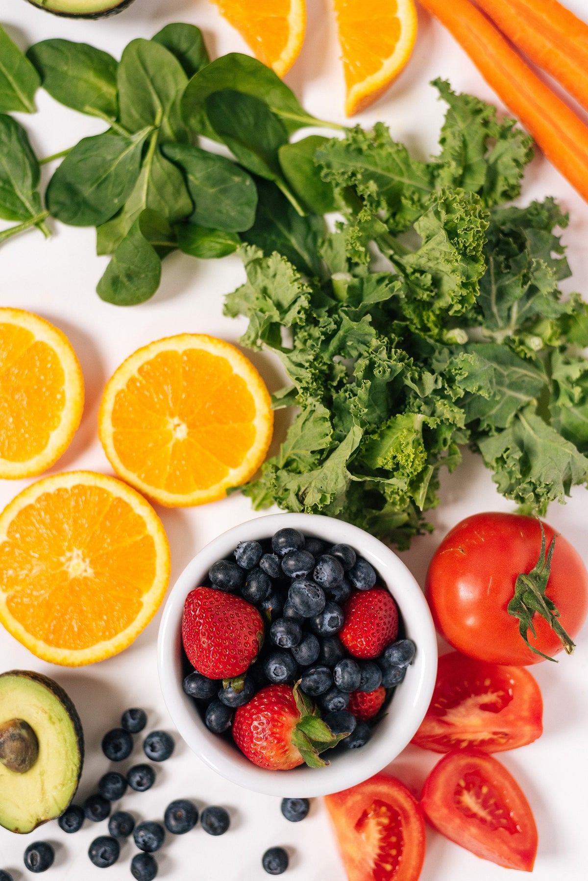 A colorful assortment of fresh fruits and vegetables, including sliced oranges, spinach, carrots, kale, a bowl of strawberries and blueberries, a whole avocado, a whole tomato, sliced tomatoes, and scattered blueberries, displayed on a white background.