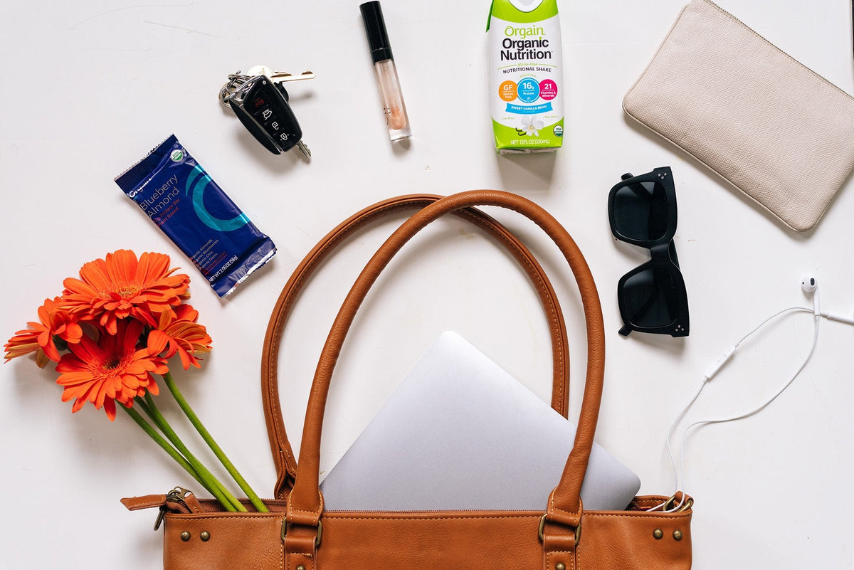 A brown leather handbag is open on a white surface with its contents displayed. Items include a laptop, sunglasses, flowers, keys, lip gloss, earbuds, a nutritional shake, a pack of gum, and a beige wallet.