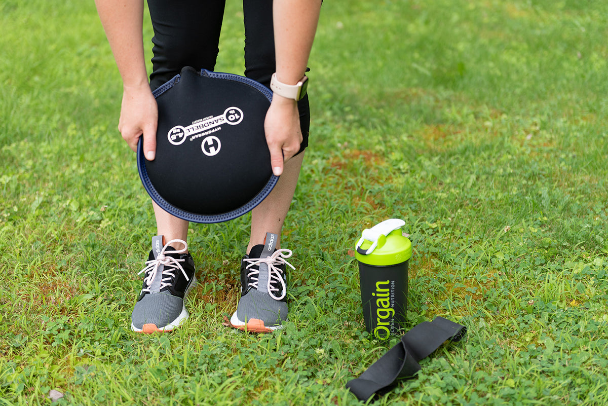 Close-up of a person bending down and holding a weight plate outdoors on grass. They are wearing sneakers, leggings, and a wristwatch. Next to them are a green shaker bottle labeled "Orgain" and a resistance band.