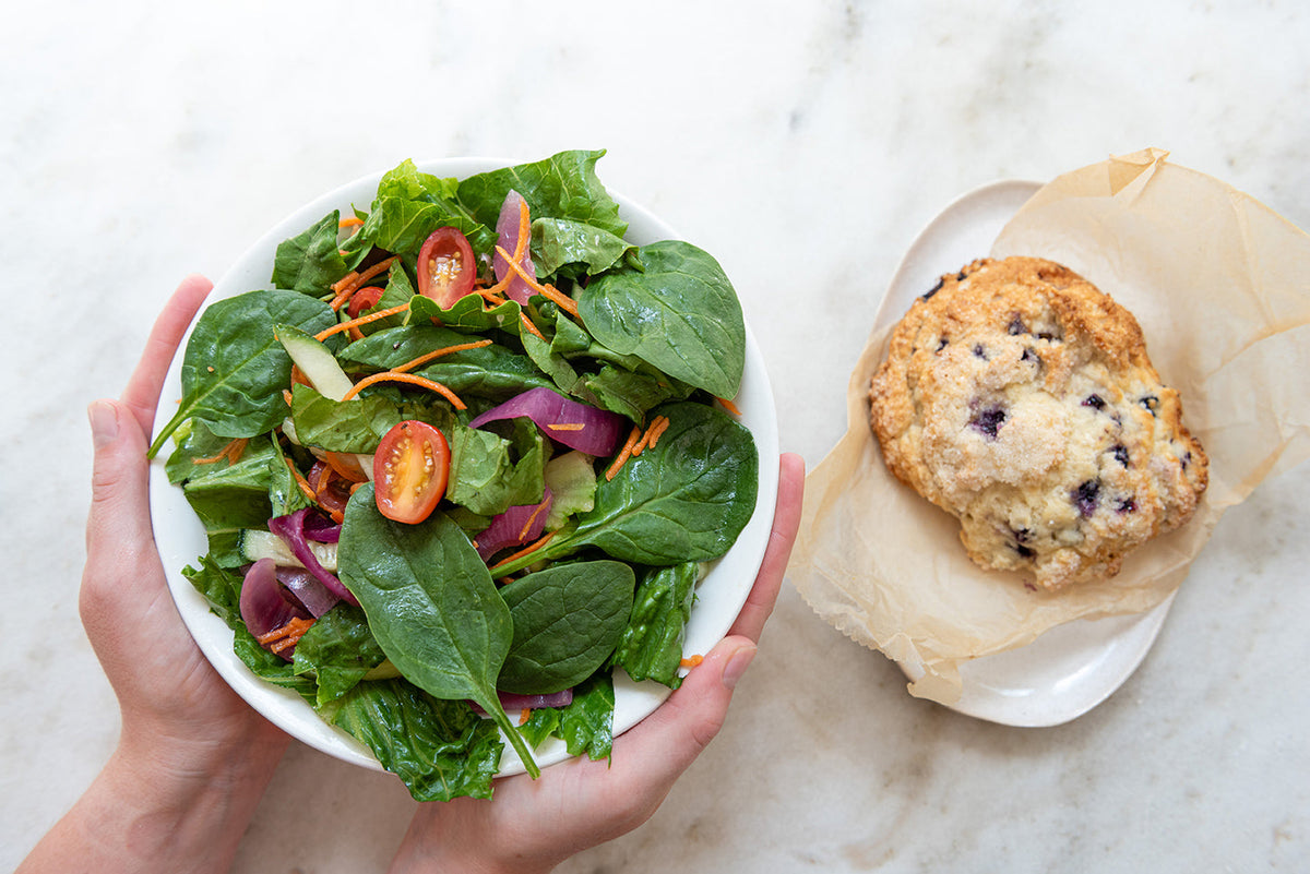 Two hands hold a bowl of fresh salad with spinach, cherry tomatoes, carrot shreds, and red onion slices next to a white plate with a blueberry scone wrapped in parchment paper on a white marble surface.