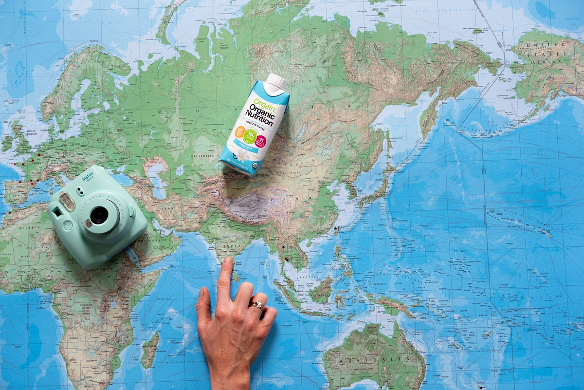 A hand pointing at a location on a world map, with a camera on the left side and a carton of organic coconut milk resting above the hand. The map shows continents of Europe, Asia, and part of Africa.