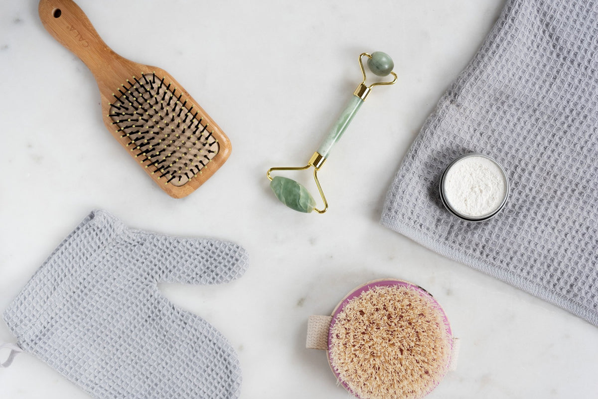 A flat lay photo of self-care items on a marble surface. Pictured are a wooden hairbrush, a gray textured towel, a jade roller, a small jar of white powder, a gray exfoliating glove, and a round loofah with a fabric handle.