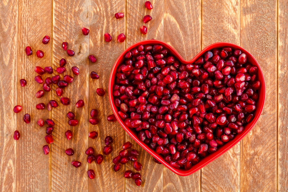 A red heart-shaped bowl filled with glistening pomegranate seeds sits on a wooden surface. Some seeds are scattered across the surface around the bowl, adding a touch of vibrant red color to the natural wood's warm tones.