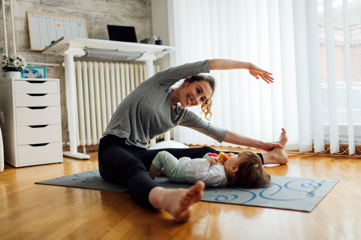 A woman stretches on a yoga mat in a living room with a baby beside her. The room has a wooden floor, white furniture, and large windows with blinds.