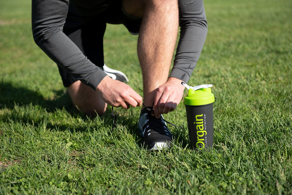Person kneeling on grass, tying the laces of black athletic shoes. Next to them is a black and green shaker bottle with the text "Orgain" visible on its side, suggesting the person is preparing for a workout or exercise session.
