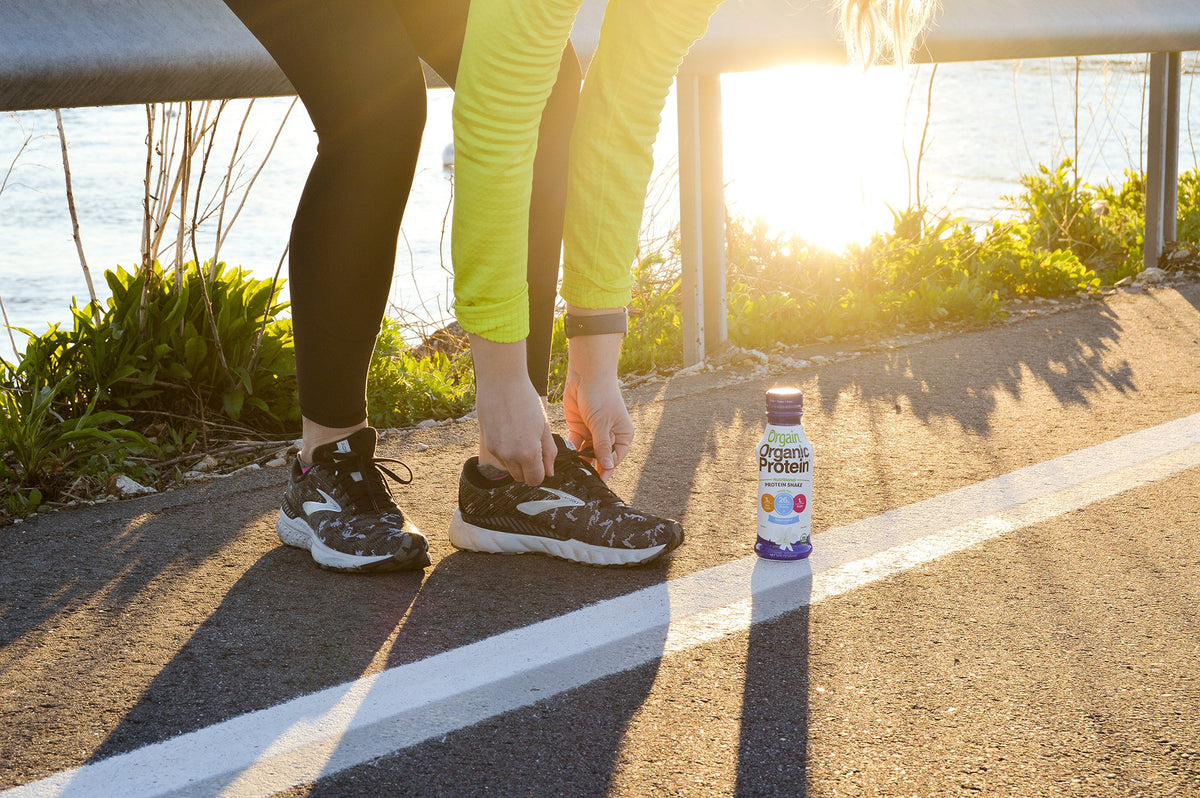 A person wearing black leggings and a bright green long-sleeve top bends down to tie their black running shoes on a paved path next to a body of water. A bottle of Orgain Organic Protein shake is placed on the ground nearby. The sun is shining brightly.