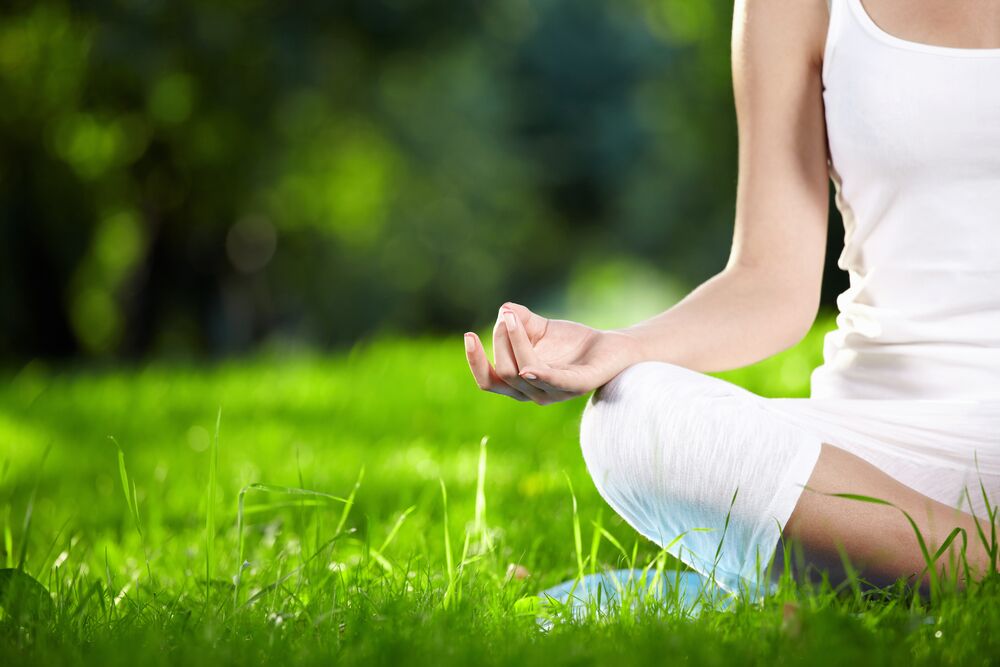 A person dressed in white sits cross-legged on a lush green lawn, meditating. Their right hand is resting on their knee, with fingers forming a mudra. The background is blurred greenery, suggesting a peaceful outdoor setting.