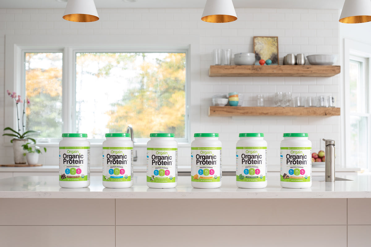 A clean, modern kitchen features a white countertop with six containers of Orgain Organic Protein powder lined up in a row. Shelves with dishes and decor are displayed in the background against a white tile backsplash. Large windows provide natural light.