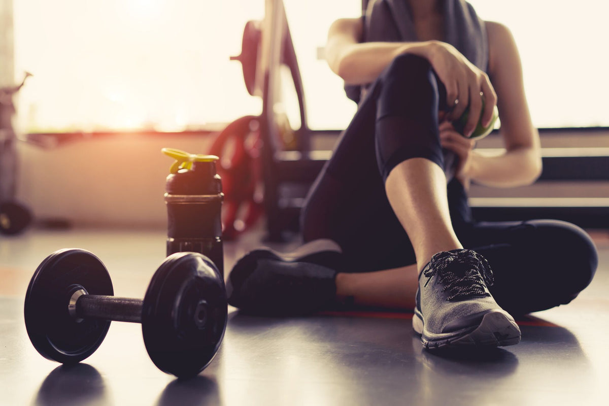 A person in athletic wear sits on the floor of a gym, resting with one leg bent and a towel around their neck. In the foreground, there is a water bottle and a dumbbell. Sunlight filters through the window, creating a warm atmosphere.