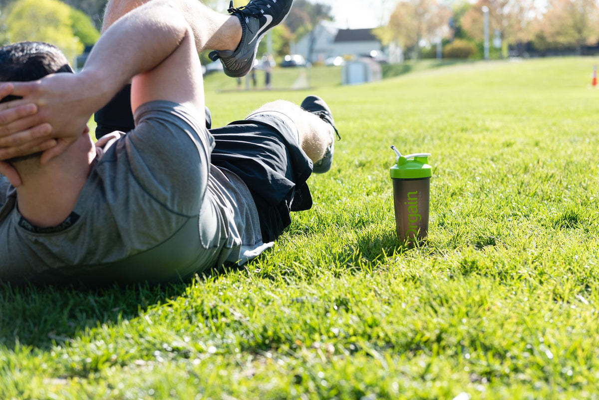 A person in athletic wear is lying on a grassy field doing sit-ups with their hands behind their head. A green bottle with a straw is next to them. The background features trees and a few buildings under a bright, sunny sky.