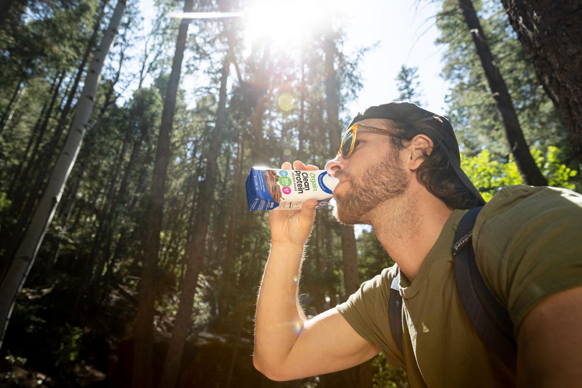 A man in a forest setting drinks from a carton of chocolate milk. Sunlight filters through tall trees, creating a serene, natural backdrop. He wears sunglasses, a cap, and a green shirt, with a backpack strap visible on his shoulder.