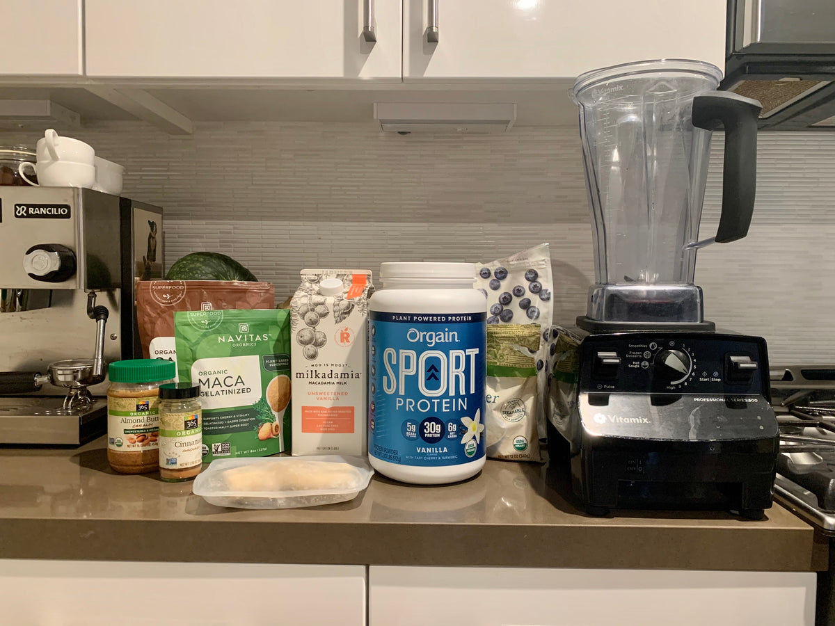 A kitchen counter with a blender and various ingredients, including a container of vanilla protein powder, a bag of macadamia nuts, ground maca powder, and a bowl of frozen banana pieces. An espresso machine and a watermelon are in the background.