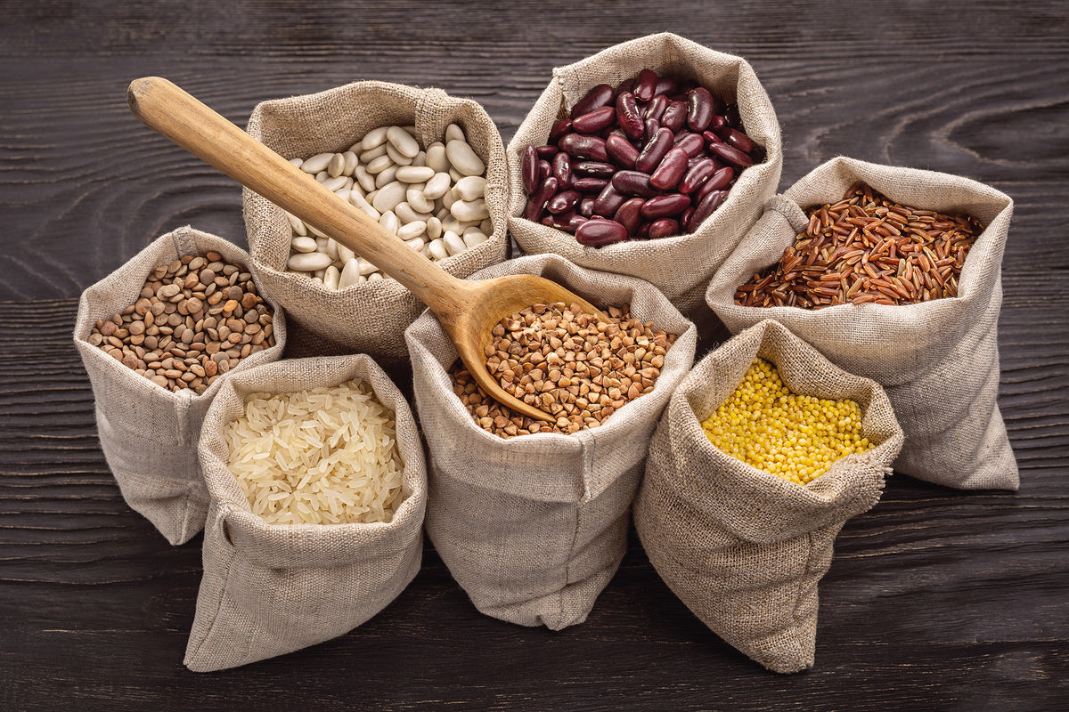 A variety of grains and legumes are displayed in small, rustic burlap sacks on a dark wooden surface. Included are beans, lentils, buckwheat, rice, red rice, and millet. A wooden spoon rests atop one sack, filled with light brown grains.