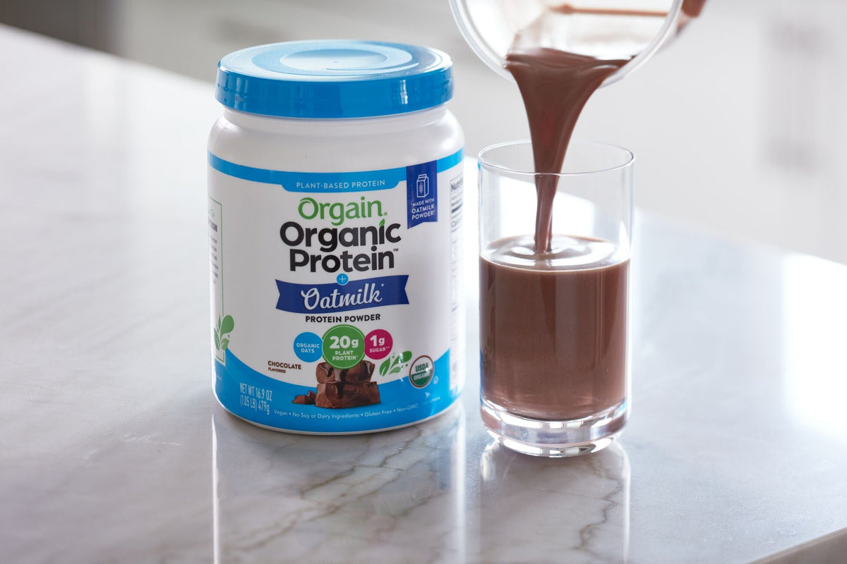 Orgain Organic Protein Powder on a counter with a glass of chocolate shake being poured. Highlights plant-based, 20g protein.