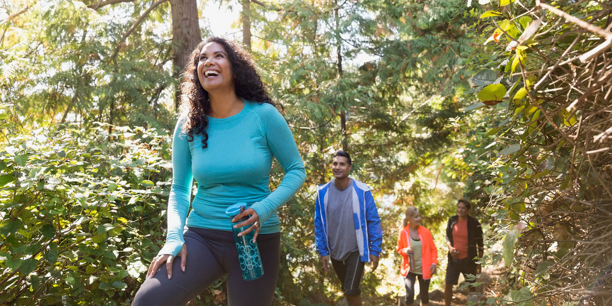 A group of people hike through a forested area on a sunny day. The woman in the foreground, wearing a blue long-sleeved shirt and holding a water bottle, smiles as she leads the group. The trail is surrounded by green trees and bushes.