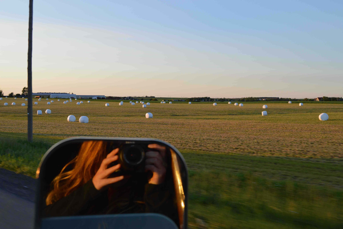 A person with long hair takes a photo with a camera reflected in a side mirror, capturing a scenic view of a field scattered with large white hay bales under a clear sky during sunset.