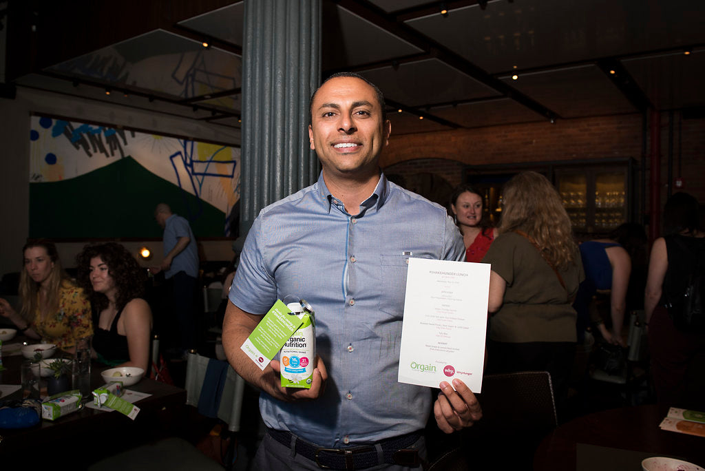 A man in a blue shirt is smiling and holding a carton of Organ organic protein almond milk and a menu. He is standing in a dimly lit restaurant with several people in the background. Some are seated at tables, engaged in conversation.