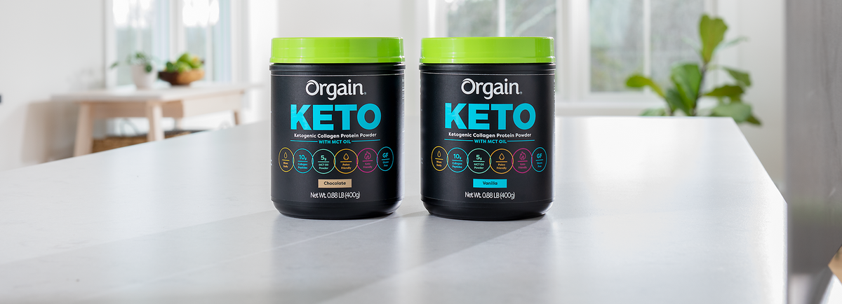 Two black containers of Orgain Keto collagen protein powder sit on a light-colored kitchen counter. One container is chocolate-flavored and the other is vanilla-flavored. The background features a bright kitchen with large windows and green plants.
