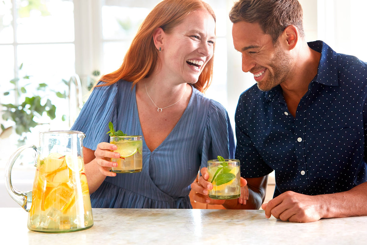 A smiling woman and man enjoy drinks at a sunny kitchen counter, with lime, mint, and a pitcher of lemon slices.