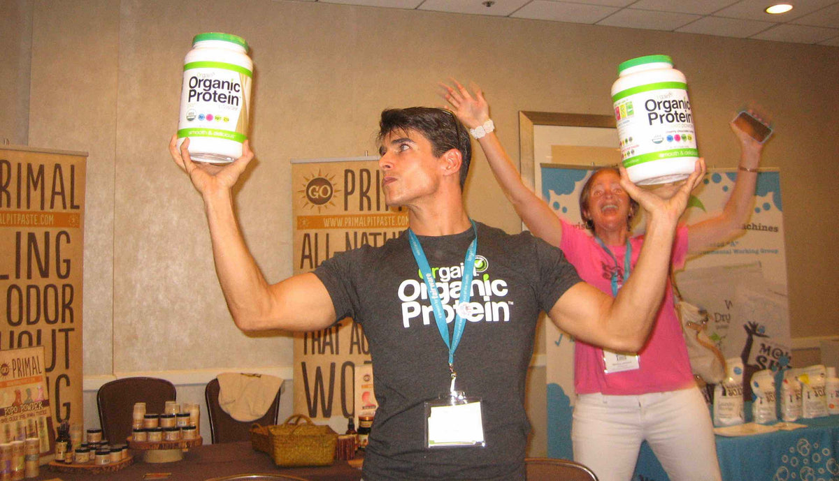 A man in a black "Organic Protein" shirt holds two large jars of organic protein powder, posing with a determined expression in front of promotional banners. A woman in the background is celebrating energetically.