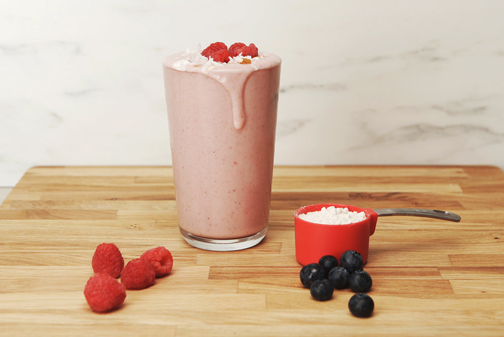 A tall glass of raspberry smoothie with a few raspberries on top, slightly overflowing, sits on a wooden surface. Nearby are several raspberries, a red measuring cup filled with a white powder, and a small cluster of blueberries.