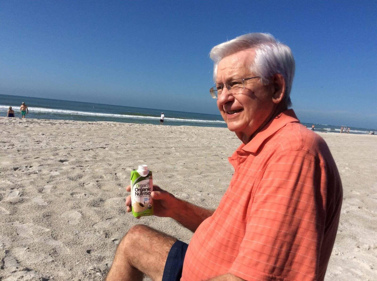 An older man with white hair, wearing an orange polo shirt and glasses, sits on a sandy beach. He is holding a carton of Organic Nutrition drink. The ocean waves and a few people are visible in the background under a clear blue sky.